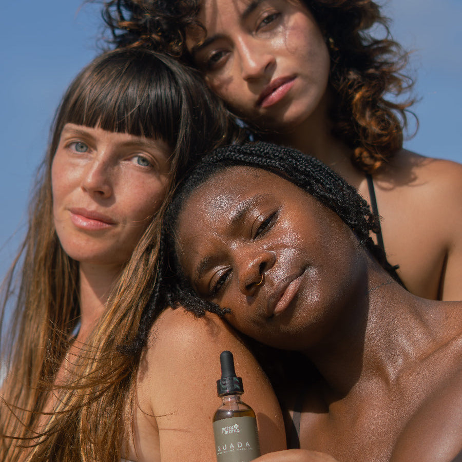 Three women standing together with a bottle of Guada nourishing hair oil against a clear blue sky.
