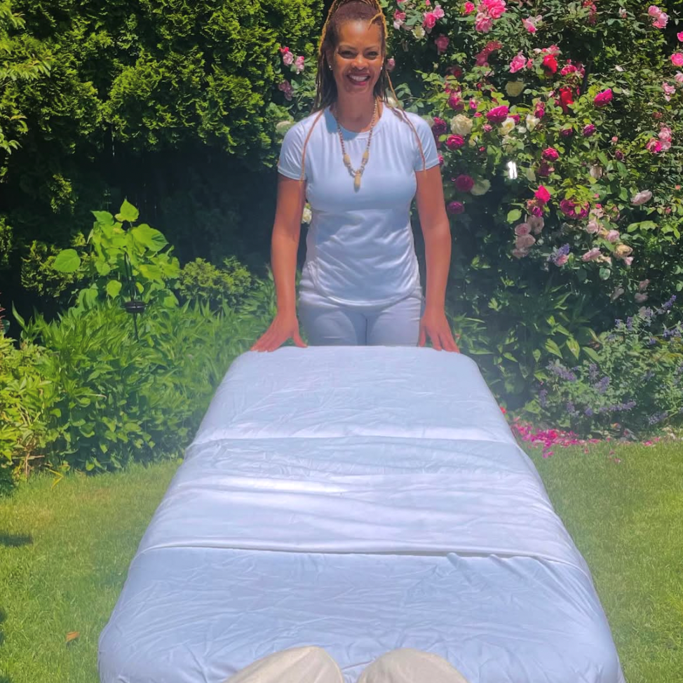 Woman standing behind a massage table outdoors with greenery and flowers in the background